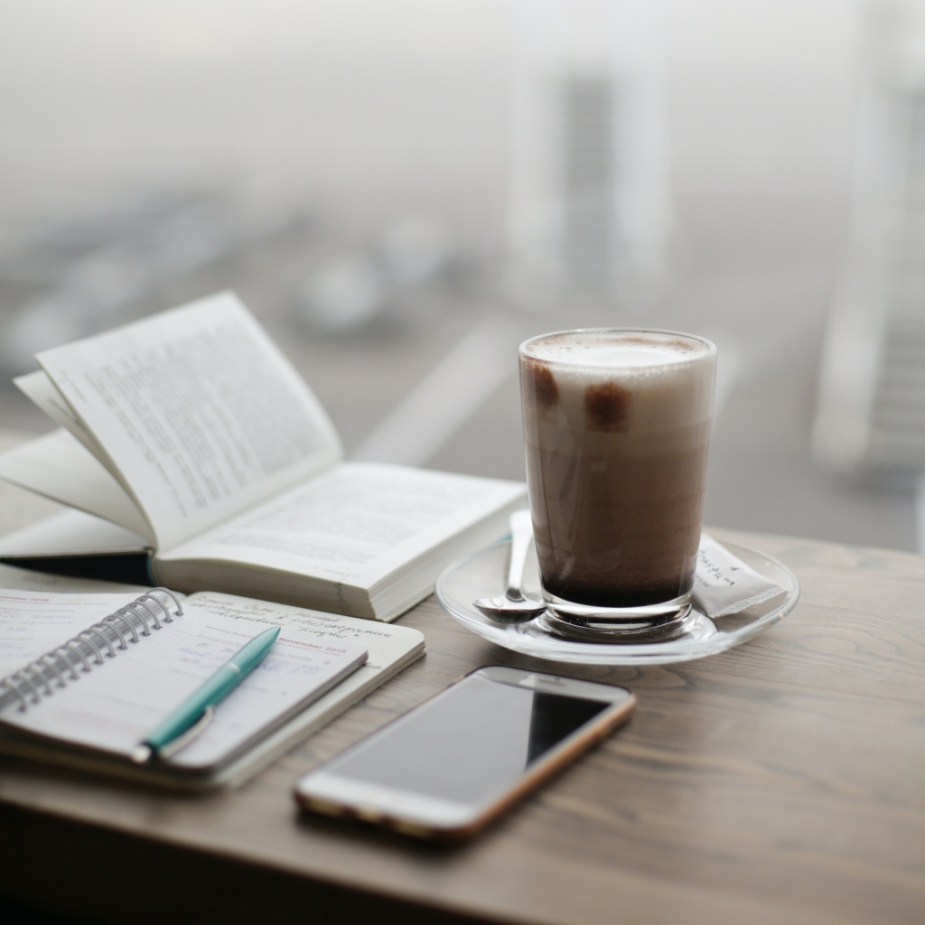 cup of coffee on a table with an open book, notebook, and phone